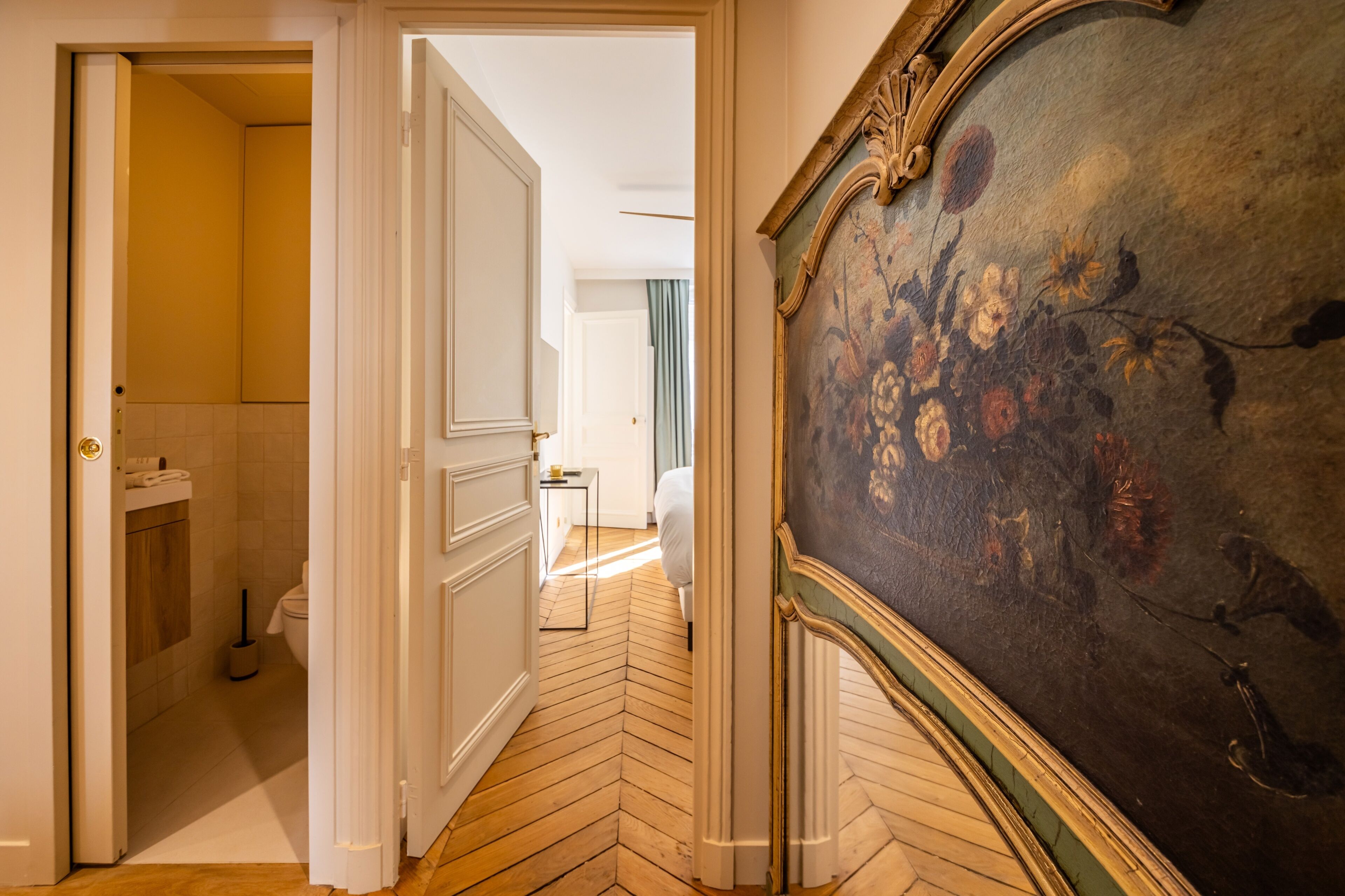 A hallway with a wooden herringbone floor, a door leading to a bathroom, and a large floral painting on the wall.