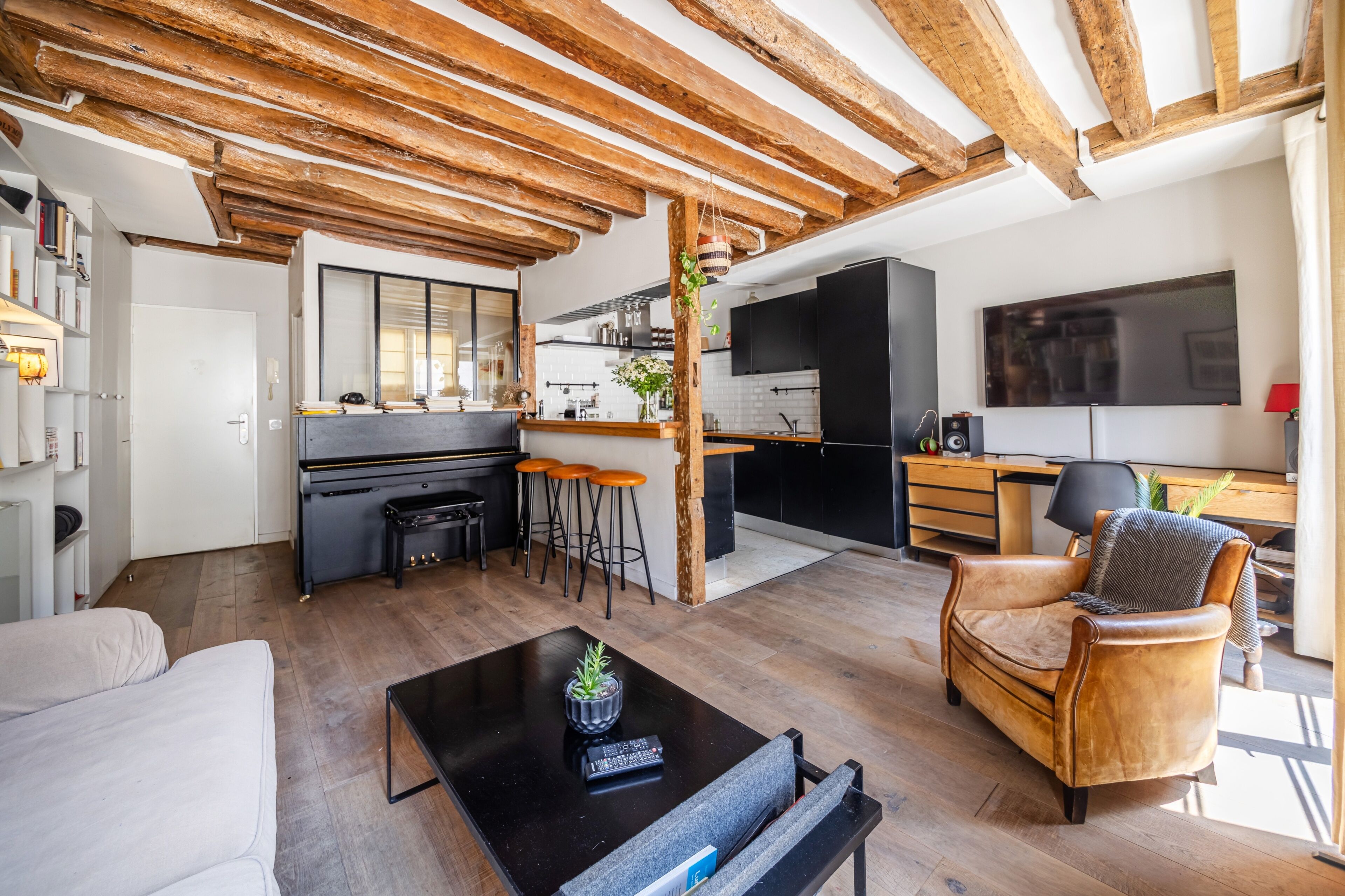 Ein moderner Wohnraum mit freiliegenden Holzbalken, einer Küche mit Barhocker, einem Sofa und einem Couchtisch mit Pflanze.