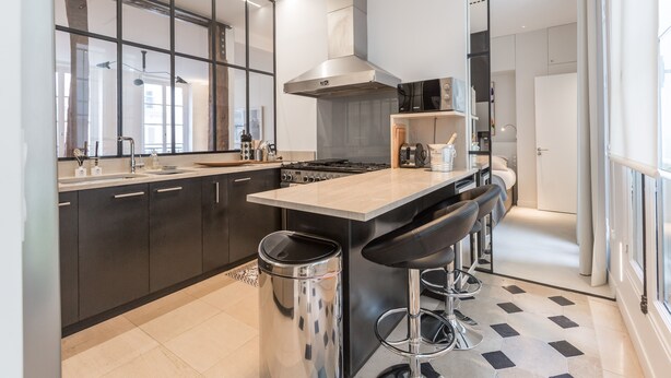 A modern kitchen with dark cabinets, a stainless steel range hood, a large island with bar stools, and a tiled floor. - Île Saint Louis Luxury (Paris)