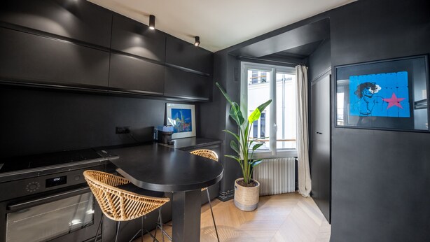A modern kitchen with dark cabinetry, a dining table, wicker chairs, a potted plant, and a framed artwork on the wall. - Opéra Style - One Bedroom Apartment, Sleeps 2 (Paris)