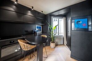 A modern kitchen with dark cabinetry, a dining table, wicker chairs, a potted plant, and a framed artwork on the wall. - Opéra Style - One Bedroom Apartment, Sleeps 2 (Paris)