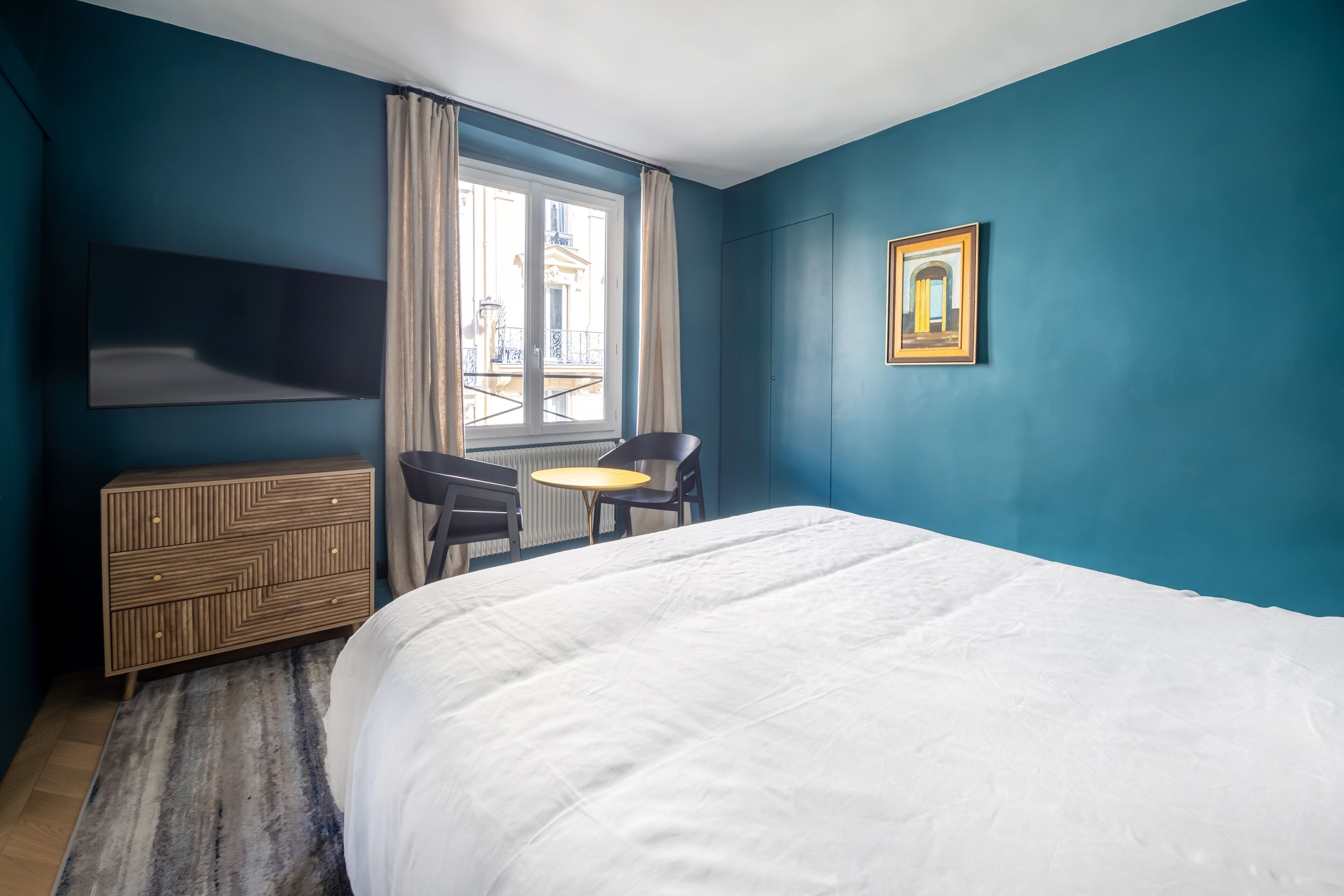 Ein Hotelzimmer mit Bett, Fernseher, Schreibtisch, Stuhl, einem Fenster mit Vorhängen und einem Bild an der Wand.