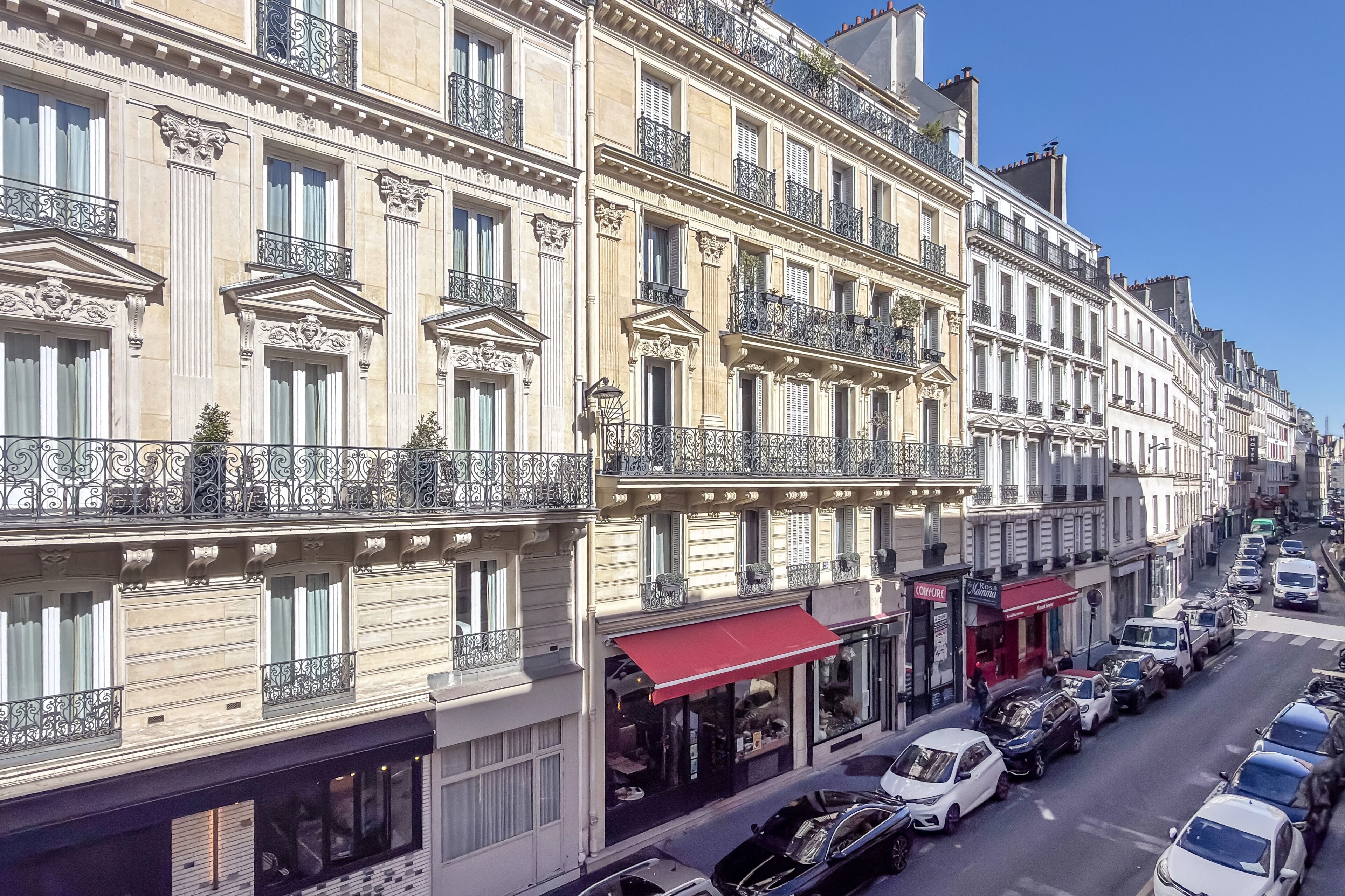 A row of classic European-style buildings with balconies, storefronts, and parked cars along a street.