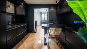 A modern kitchen with dark cabinetry, a central island with chairs, a large green plant, and a view to a balcony. - Opéra Style - One Bedroom Apartment, Sleeps 2 (Paris)