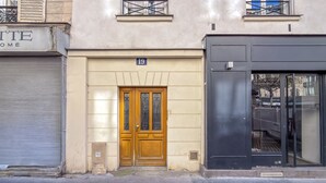 A building with a closed shutter on the left, a wooden door with a number 19 sign above it, and a black storefront on the right. - Opéra Style - One Bedroom Apartment, Sleeps 2 (Paris)