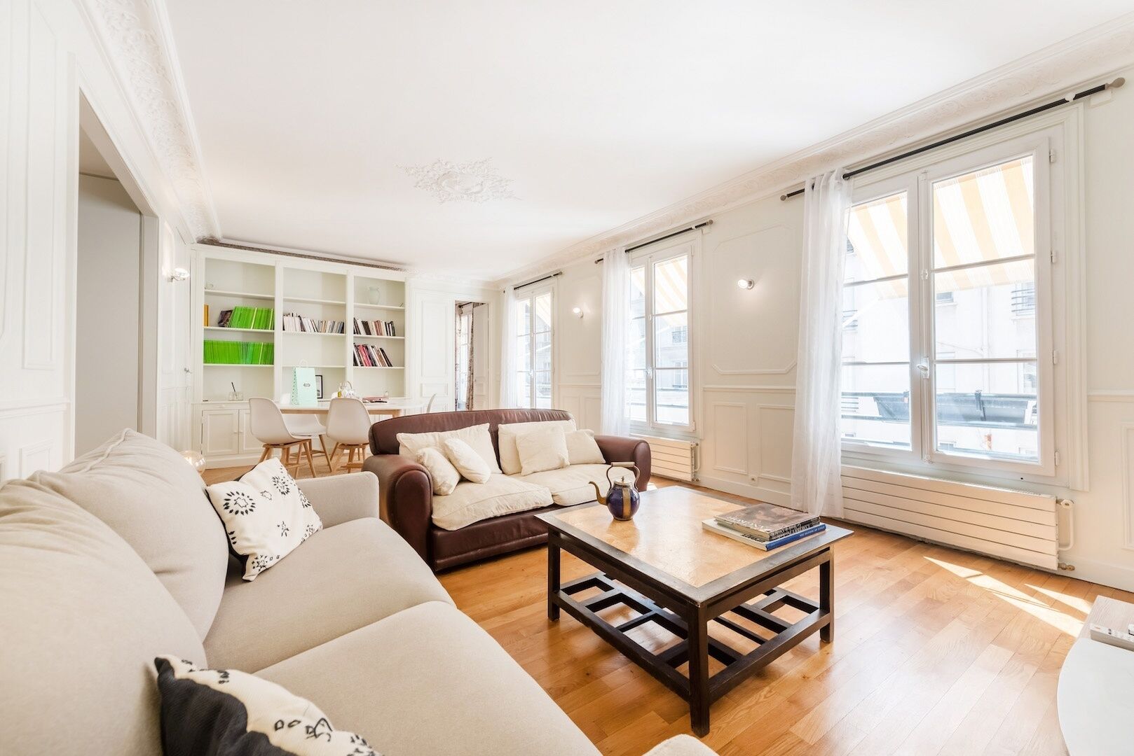 A living room with a brown sofa, a beige sofa, and a coffee table. There is a bookshelf with books and a green box.