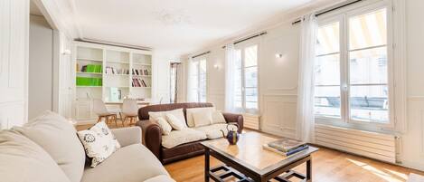 A living room with a brown sofa, a beige sofa, and a coffee table. There is a bookshelf with books and a green box.