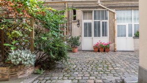 A courtyard with a cobblestone path, potted plants, a wooden lattice, and a building with a white door and windows. - Bon Marché Sevres - Two Bedroom Apartment, Sleeps 4 (Paris)