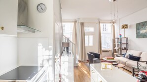 A modern kitchen with a stove, a dining area with a table and chairs, a sofa, and a bookshelf. - Bon Marché Sevres - Two Bedroom Apartment, Sleeps 4 (Paris)