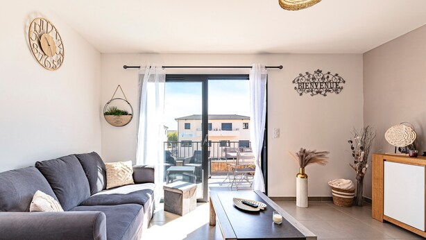 Una sala de estar moderna con un sofá gris, una mesa de centro, un reloj de pared, un cartel de bienvenida y vistas al exterior.