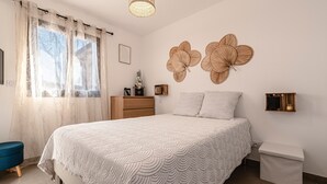 Un dormitorio con cama, mesitas de noche, cómoda, ventana con cortinas translúcidas y elementos decorativos en la pared.