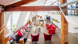 Un soggiorno in stile loft con soffitto alto, travi in legno, una divano, poltrone rosse, un tavolino e una cucina sullo sfondo.
