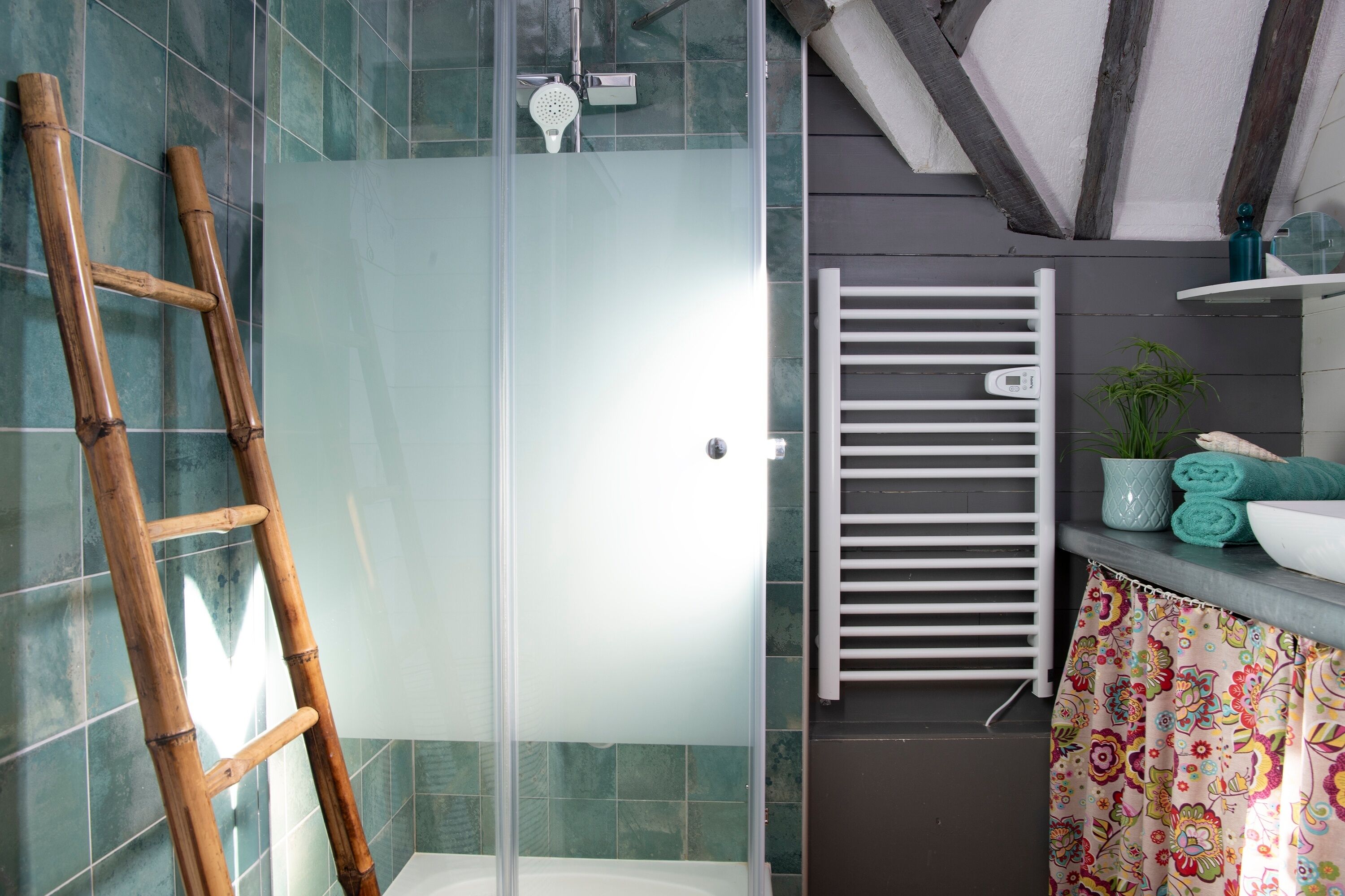 A bathroom with a glass shower enclosure, a white towel rack, a patterned shower curtain, and a bamboo ladder.