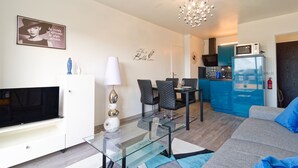 Ein modernes Wohnzimmer mit Flachbildfernseher, Glastisch und Sofa. Es gibt einen Essbereich mit Tisch und Stühlen, eine Küche mit Mikrowelle und Kühlschrank, sowie eine Kristallleuchter.