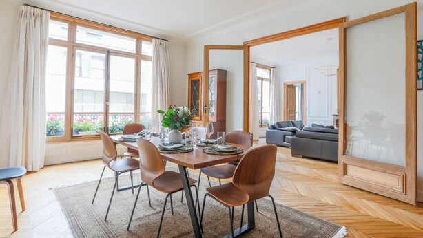 A dining area with a table set for four, surrounded by chairs, with a view of the outside through large windows. - Jardin du Luxembourg Home (Paris)