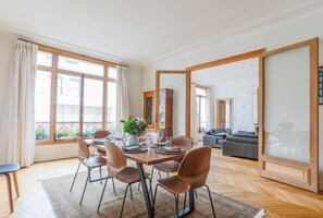A dining area with a table set for four, surrounded by chairs, with a view of the outside through large windows. - Jardin du Luxembourg Home (Paris)