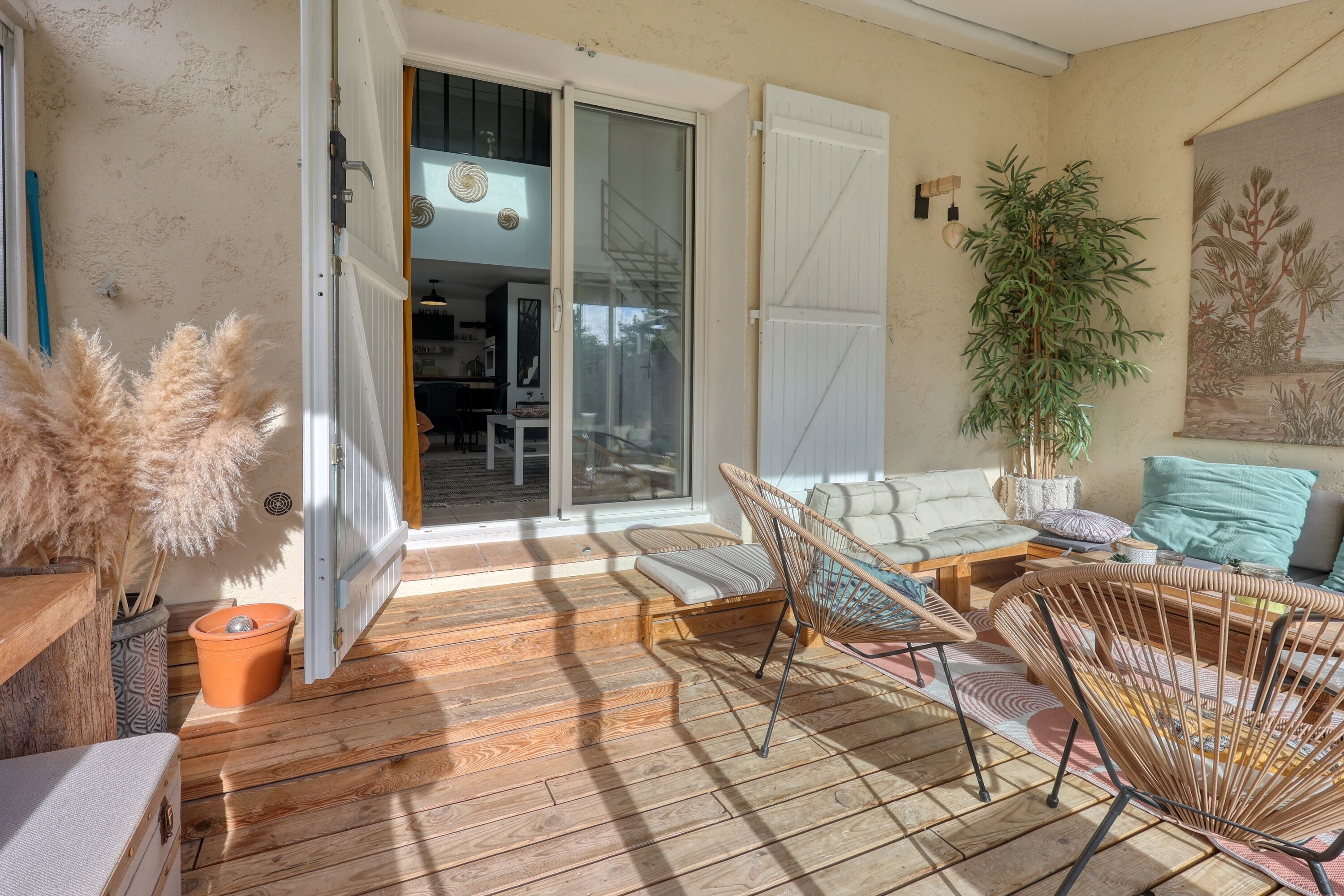 Une porte coulissante ouverte donne sur un balcon doté d’une terrasse en bois, de chaises en osier, d’une plante en pot et d’un tableau accroché au mur.