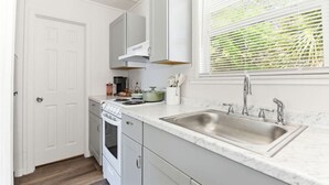 Une cuisine avec des meubles blancs, un évier en acier inoxydable, une cuisinière et une fenêtre avec des stores.