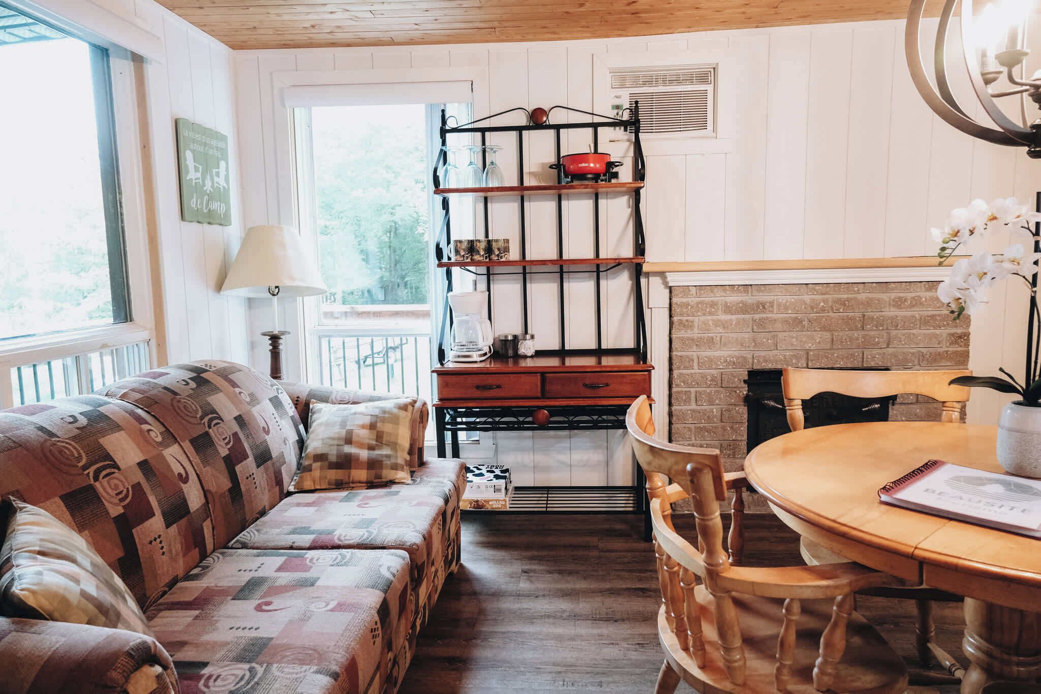 A cozy room with a wooden ceiling, a brick fireplace, a wooden dining table with chairs, a patterned sofa, and a metal shelf with kitchen items.