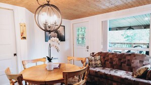 A wooden dining table with chairs, a chandelier, a patterned sofa, and a window with a view of trees. - Nature Escape and Miller Zoo | Buddy Shack (Frampton)