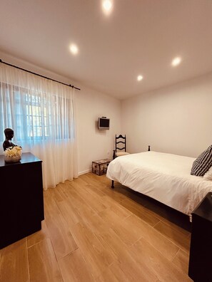 Une chambre à coucher avec un lit, une commode, un téléviseur fixé au mur et une fenêtre avec des rideaux.