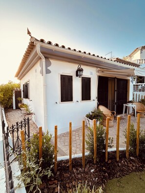 Un bâtiment blanc avec un toit en tuiles, des volets noirs et un petit porche meublé de meubles en osier.