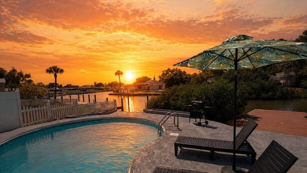 Ein Schwimmbad mit Liegestühlen und einem Sonnenschirm, umgeben von einer Terrasse und Blick auf den Sonnenuntergang.