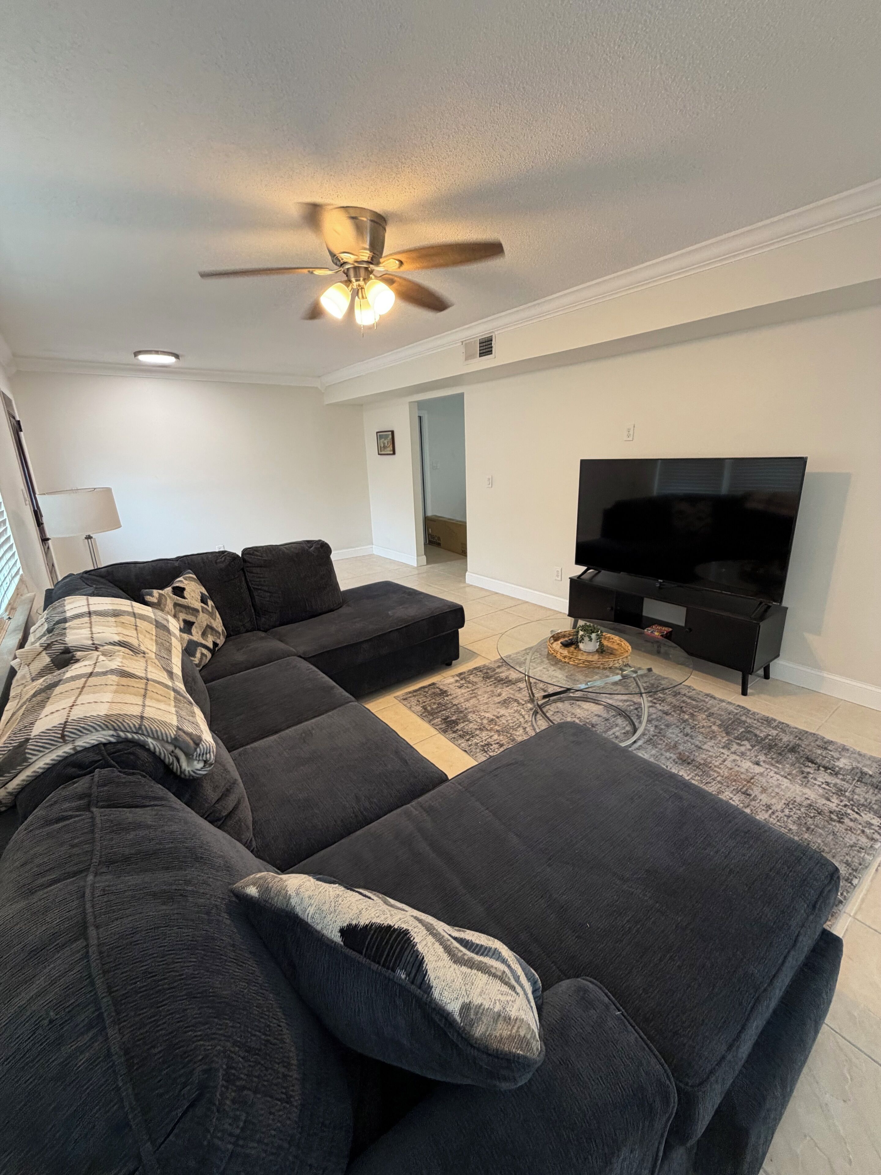 A living room with a sectional sofa, a flat-screen TV, a ceiling fan, and a lamp.