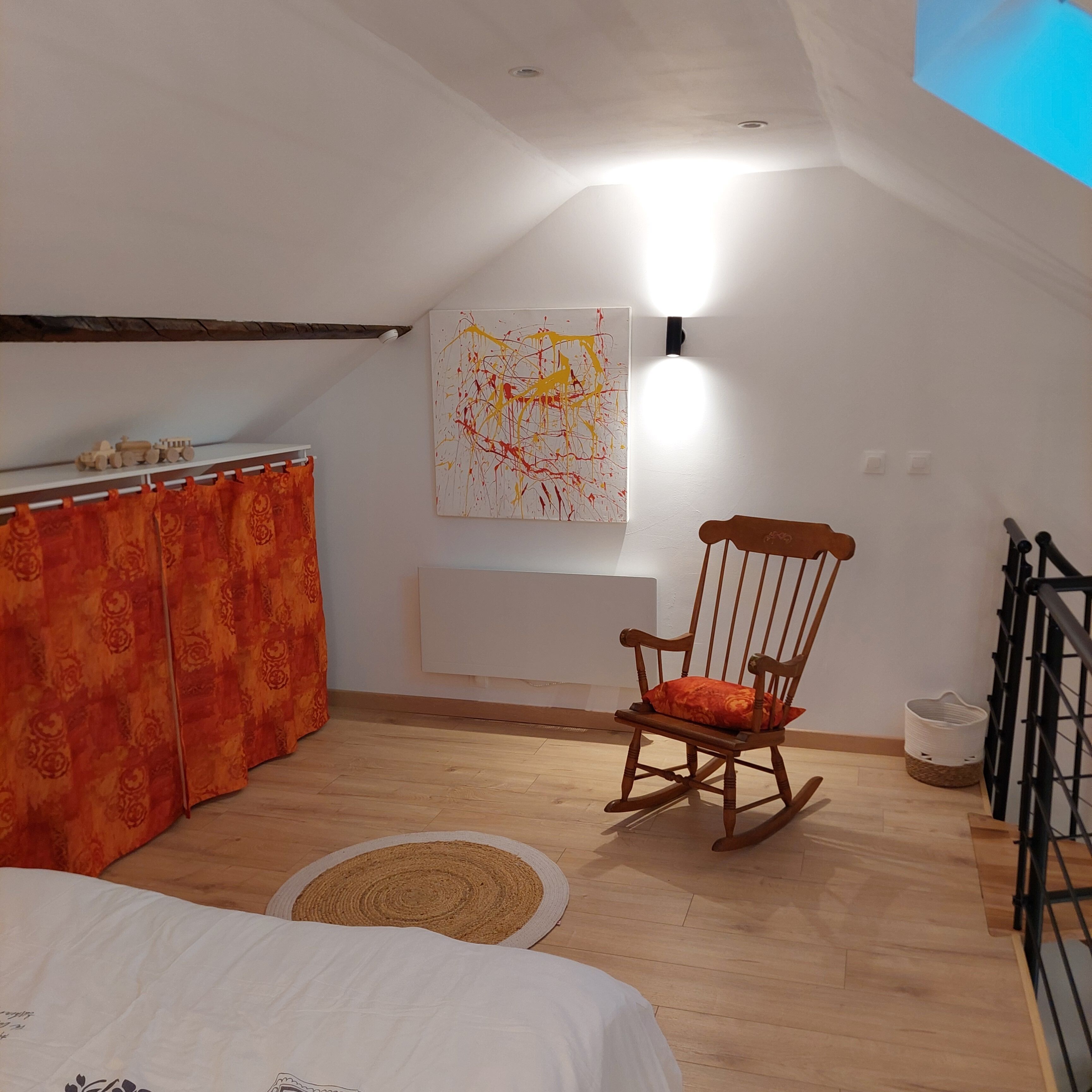 Een kamer met een houten schommelstoel, een bed met wit beddengoed, een rood gordijn met patroon, een witte muur met een schilderij en een dakraam.