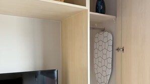 Et værelse med et fjernsyn på et hvidt stativ, et lille bord med et lys, og et klædeskab med et håndklæde med geometrisk mønster hængende.