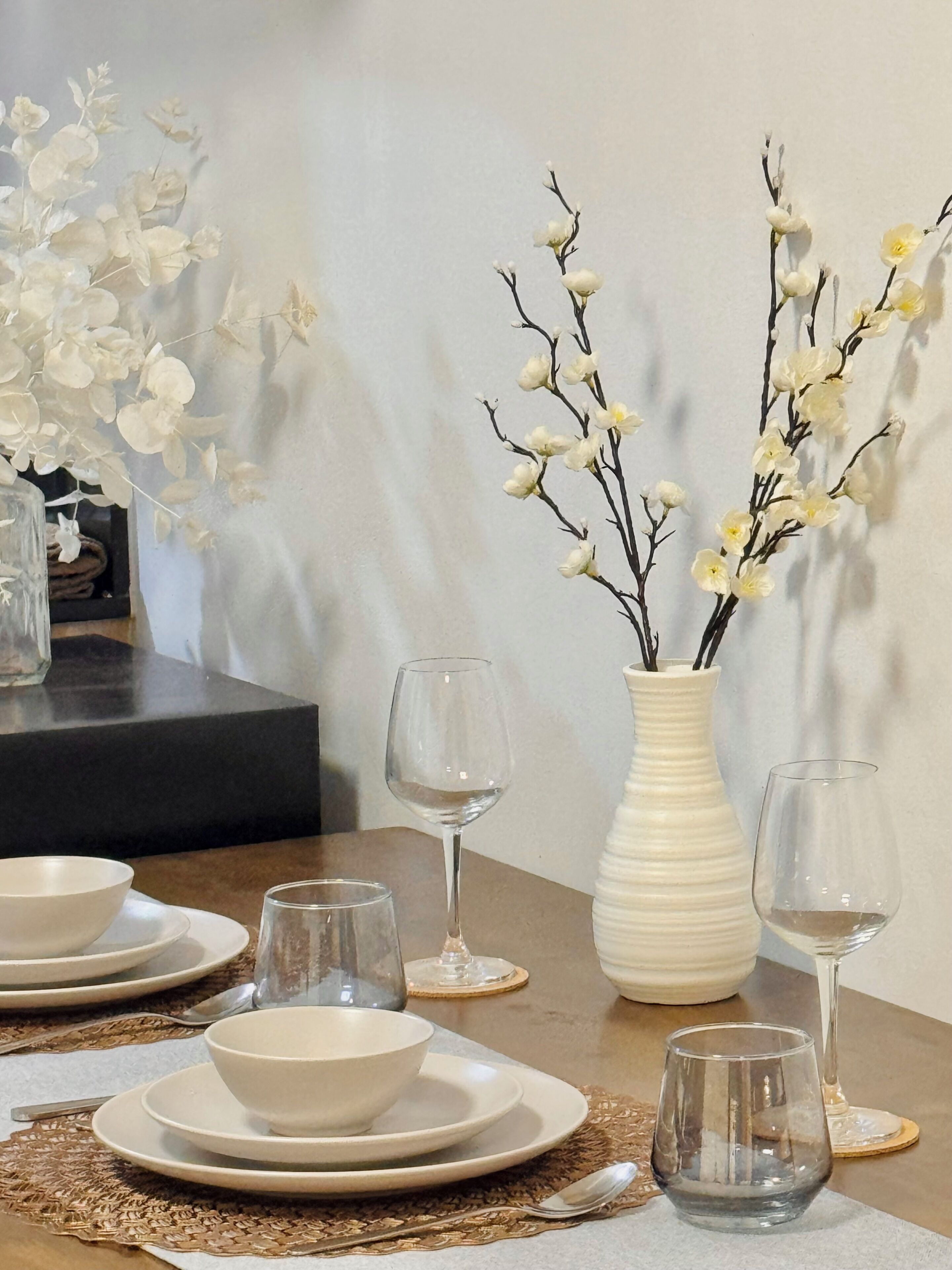 A dining table set with plates, bowls, and wine glasses, accompanied by a vase with branches and flowers.