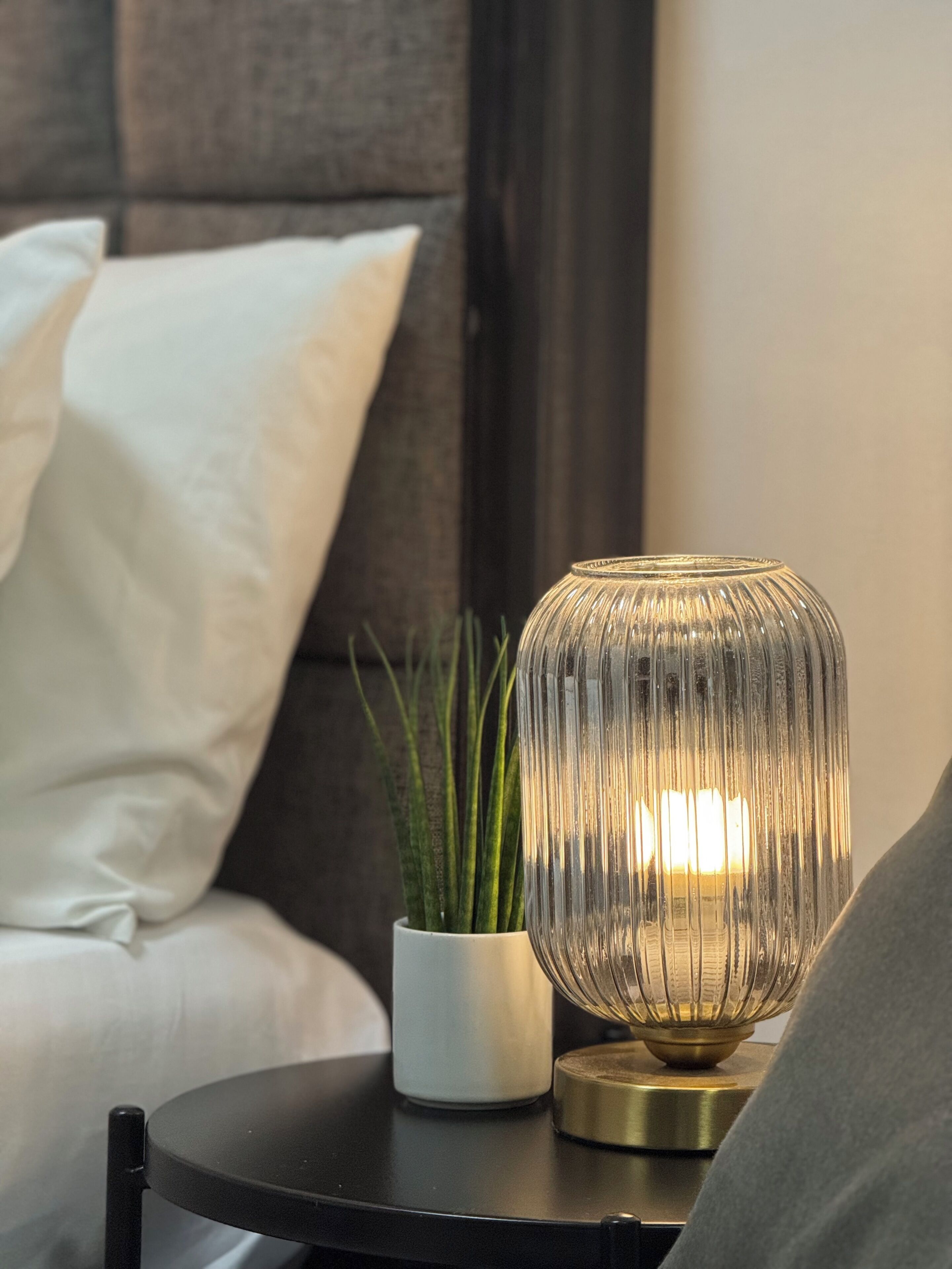 A bedside table with a clear glass lamp, a white vase with green plants, and a white pillow on a dark-colored bed.