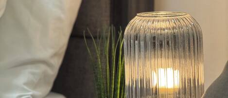 A bedside table with a clear glass lamp, a white vase with green plants, and a white pillow on a dark-colored bed.