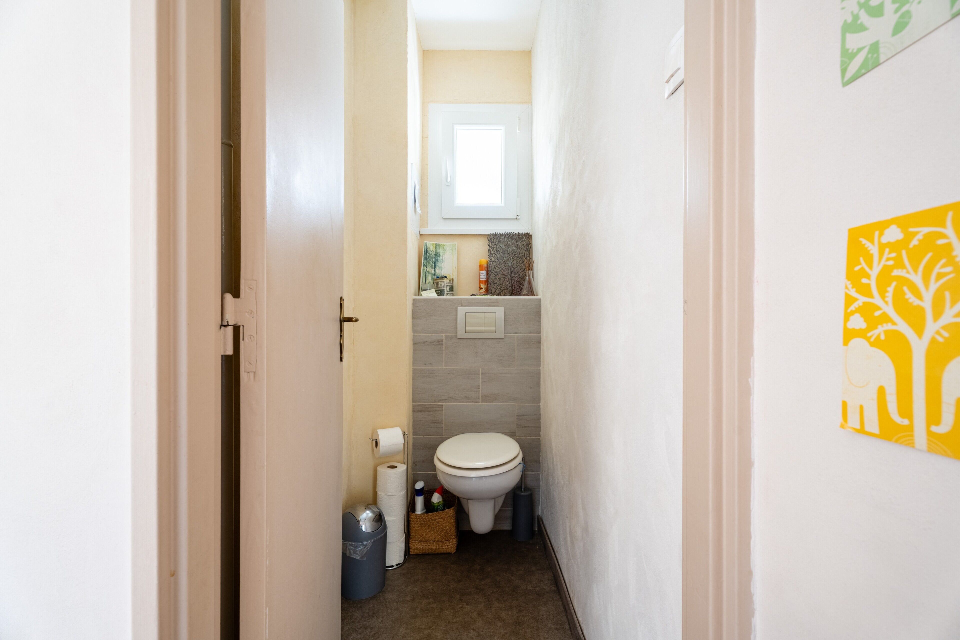 A narrow hallway leading to a bathroom with a toilet, a trash bin, and a basket of toiletries.