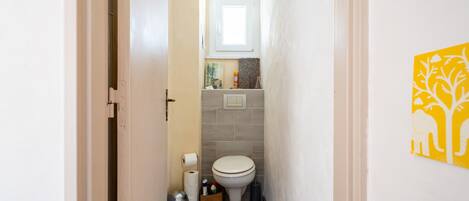A narrow hallway leading to a bathroom with a toilet, a trash bin, and a basket of toiletries.