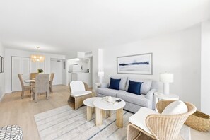 Een moderne woonkamer met een grijze bank, witte stoelen, een salontafel en een eethoek met een tafel en stoelen.