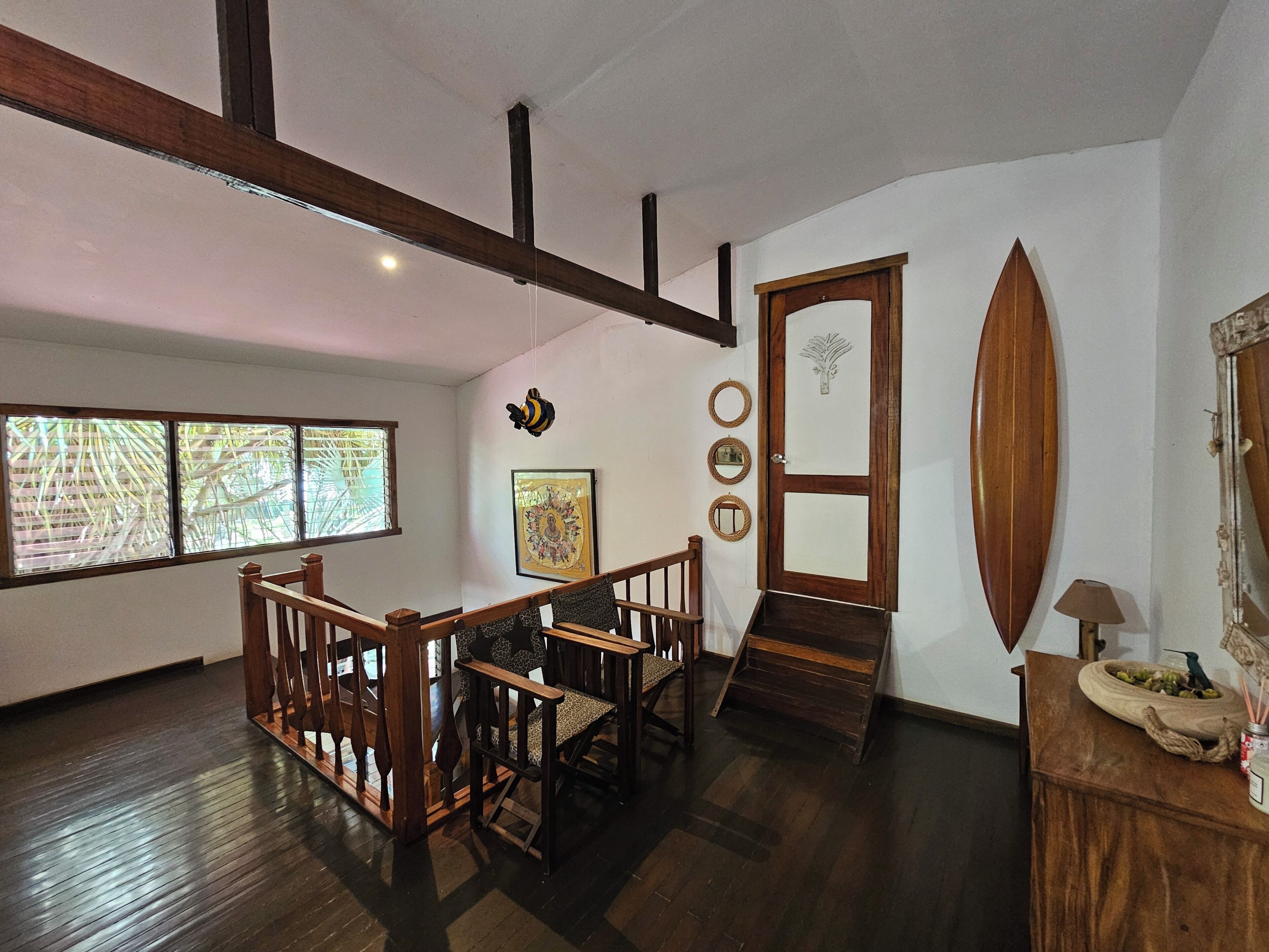 Une pièce dotée d’un escalier en bois, d’un coin repas avec une table et des chaises en bois, d’une grande fenêtre avec des stores et d’une porte ornée d’un miroir et d’objets décoratifs.