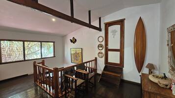 Une pièce dotée d’un escalier en bois, d’un coin repas avec une table et des chaises en bois, d’une grande fenêtre avec des stores et d’une porte ornée d’un miroir et d’objets décoratifs.