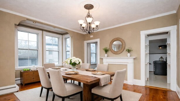 A dining room with a wooden table, six chairs, a chandelier, a fireplace, and large windows.
