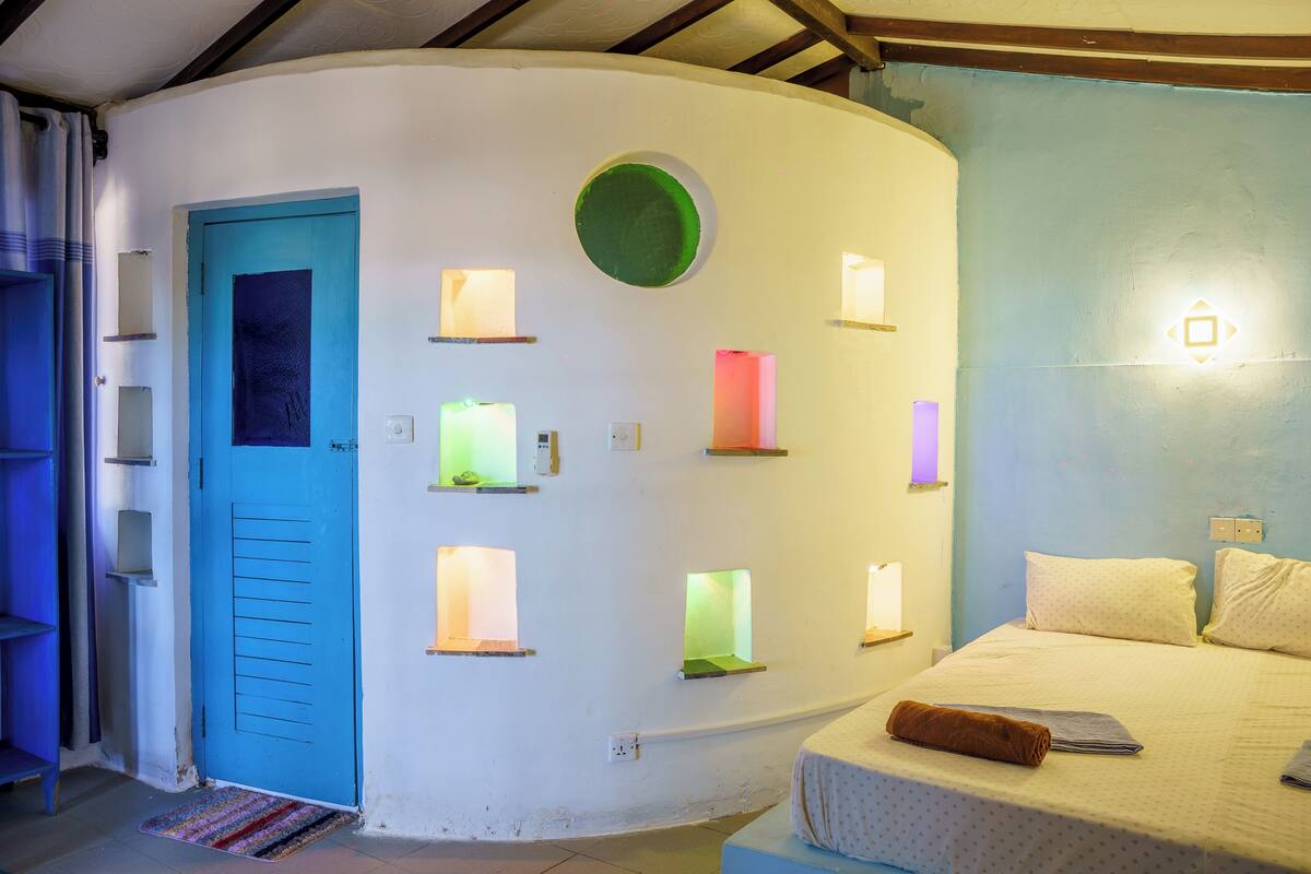 Una habitación con una puerta azul, una cama con cubrecama blanco y varias lámparas fijadas a la pared.