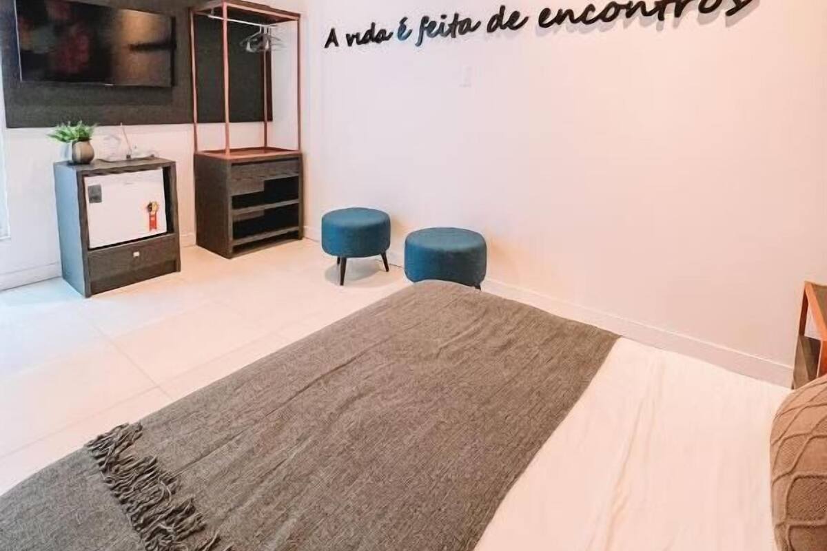 A bedroom with a bed, a wall-mounted TV, a small desk, two blue ottomans, and a motivational quote on the wall.