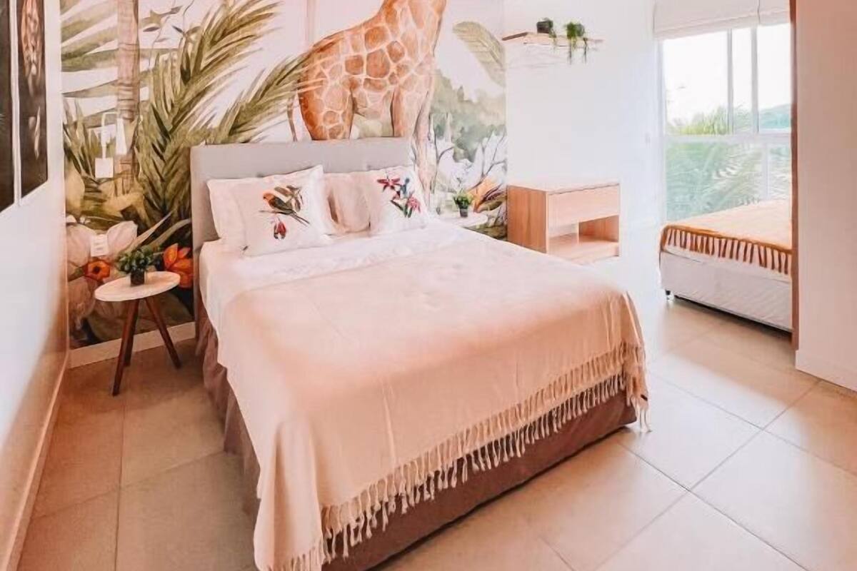 A bedroom with a bed, a nightstand, a window, and a mural of a giraffe and a parrot.