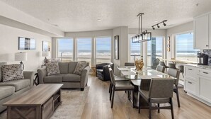 A spacious living area with a large window overlooking the beach, a dining area with a glass table, and a kitchen with white cabinets. - Promenade 403 - Oceanfront Oasis - Magical Views (Seaside)