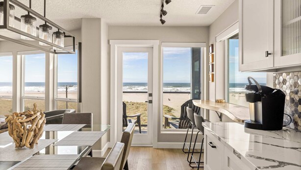 A modern kitchen with a view of the beach, a dining area with a glass table, and a bar area with a coffee machine. - Promenade 403 - Oceanfront Oasis - Magical Views (Seaside)