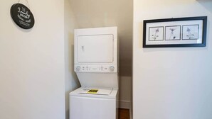 Un cuarto de lavado con lavadora y secadora, un letrero que dice 'The Laundry Room', y un cuadro con flores en la pared.