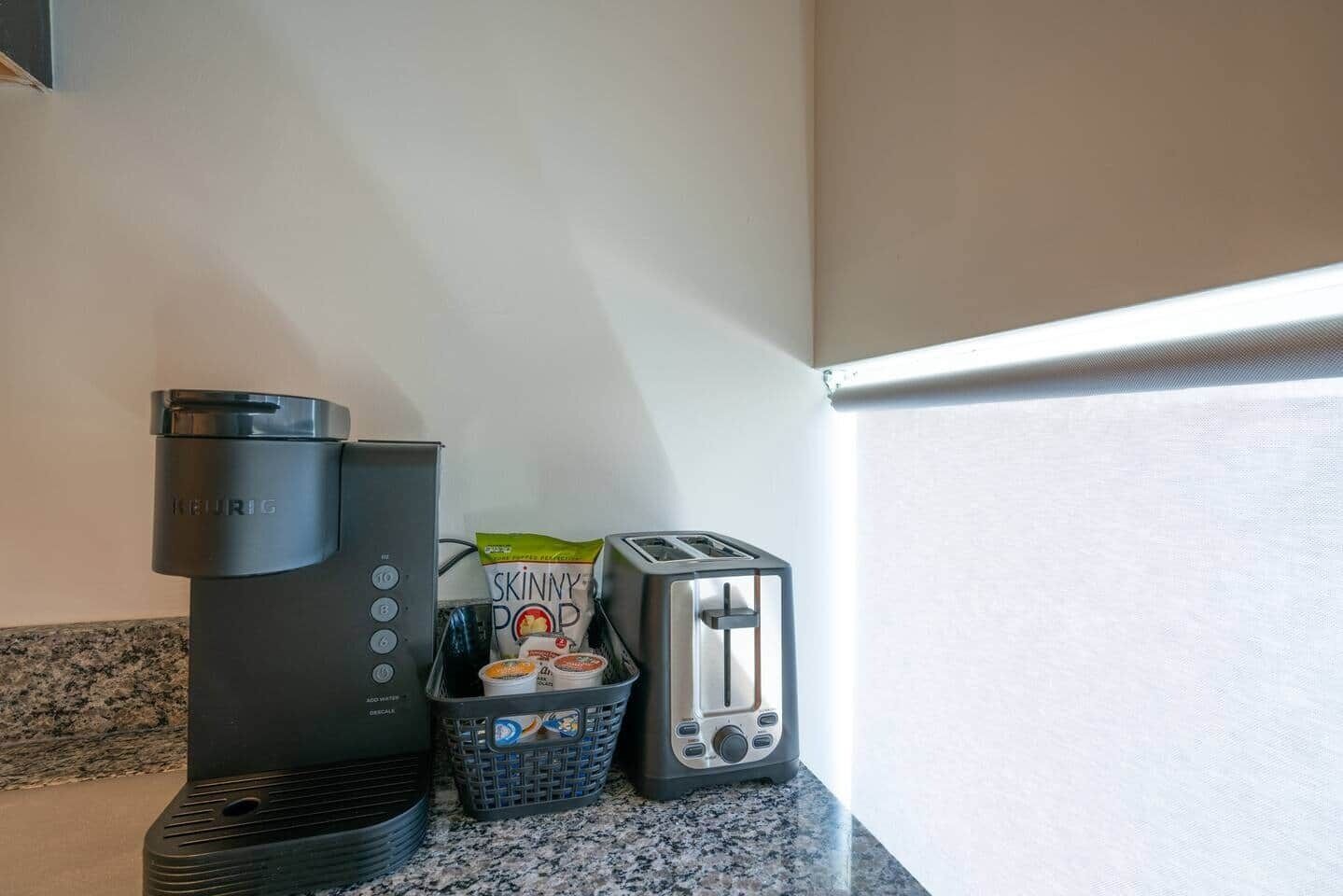 A coffee maker, a toaster, a basket with snacks, and a bag of Skinny Pop on a kitchen counter.