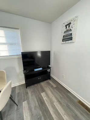 Una habitación con una televisión de pantalla plana sobre un soporte negro, una silla blanca, una ventana con persianas y una pared con un cartel.