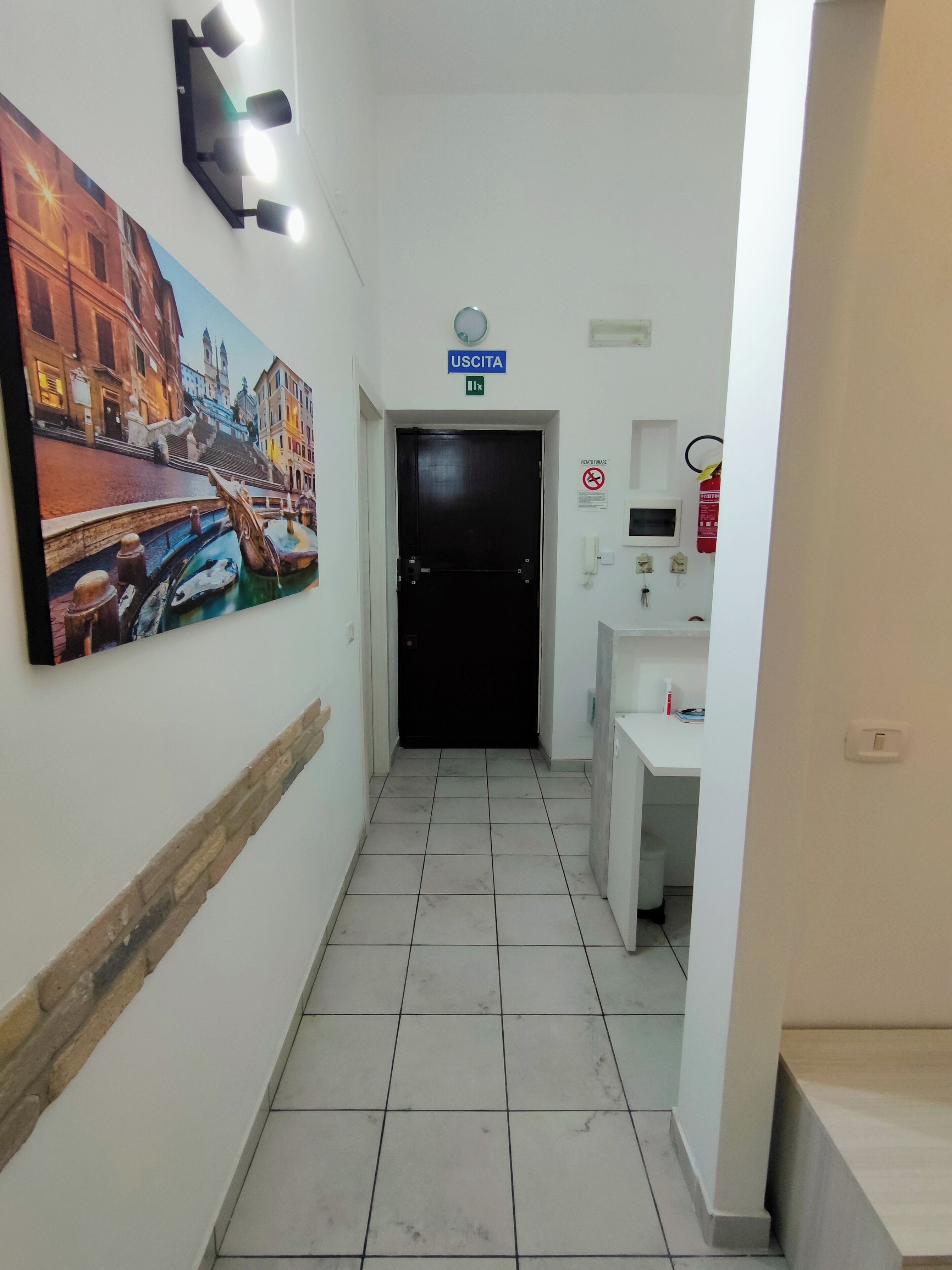 A hallway with a tiled floor, a black door, a 'USCITA' sign, a fire extinguisher, and a wall-mounted light fixture.