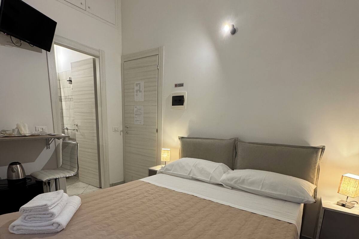 A hotel room with a bed, a TV mounted on the wall, a bedside table with a lamp, and a bathroom visible through an open door.