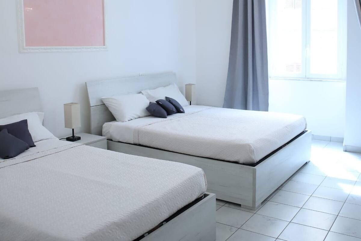 A modern hotel room with two beds, a large window with curtains, and a minimalist design.